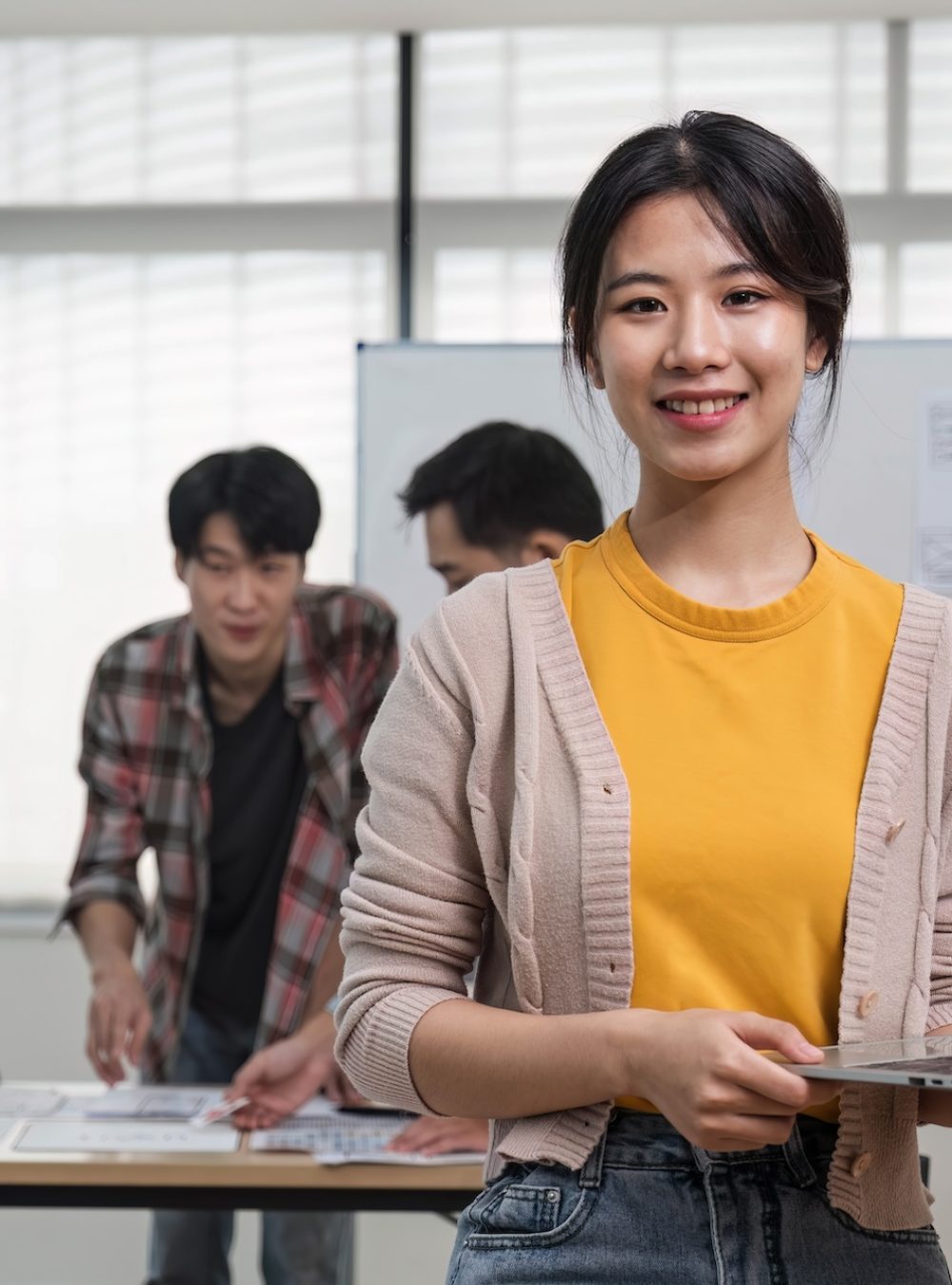 young Asian female marketing assistant is working on her work on her laptop at her desk while her coworkers are working in the room